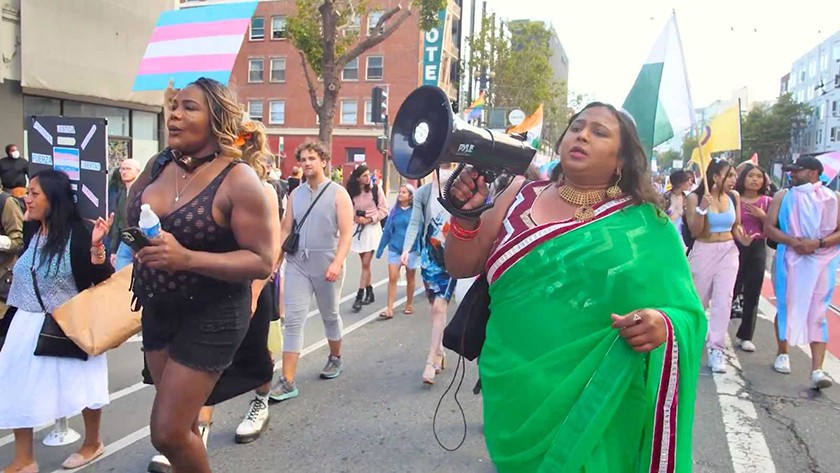 A parade holding megaphones and trans flags marches down a street