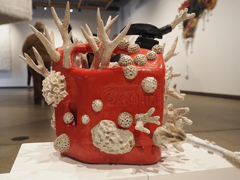 A gallery shows people in the background and a multi-media piece in the foreground. The artwork mimics a red gasoline can, accessorized by tan fossil-like objects