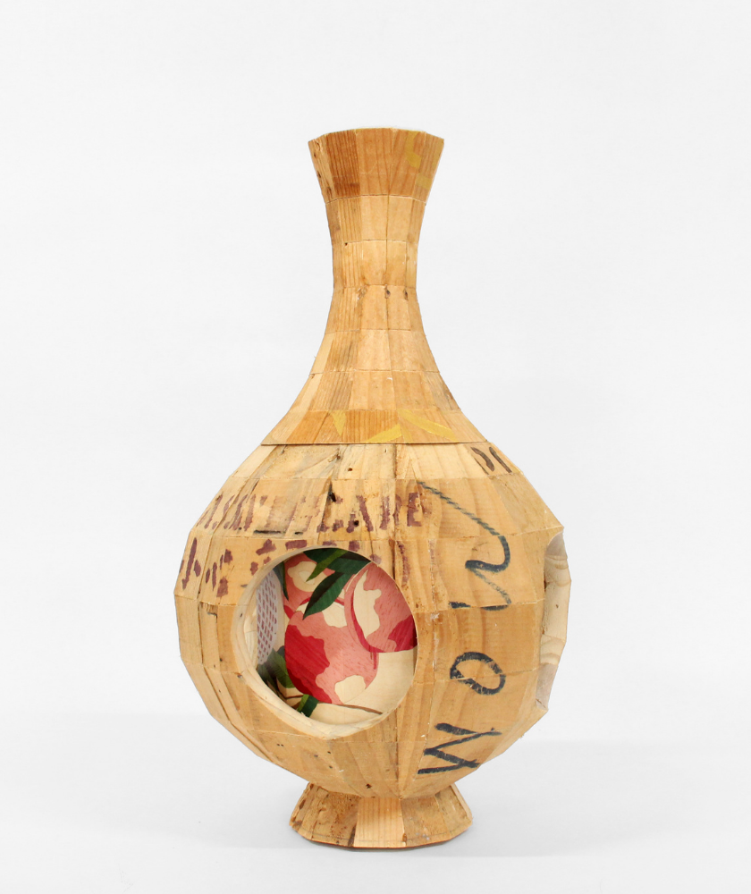 A wooden vase-shaped sculpture with a circular cutout, revealing a colorful floral-patterned object inside. The surface of the vase features visible seams and handwritten text. The background is plain white.