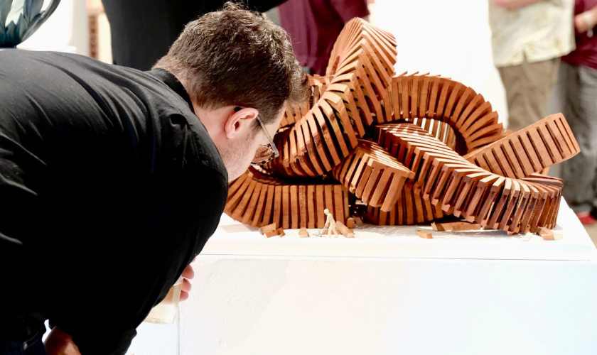 A person closely observes an intricate wooden sculpture made of interlocking rectangular pieces, displayed on a white pedestal in an art gallery. Other visitors are visible in the blurred background.