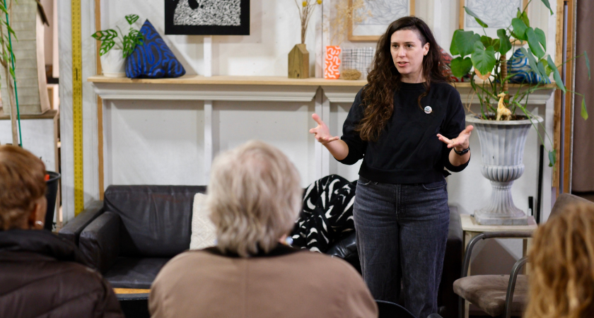 A woman with long brown hair speaks to a group of people inside an art space
