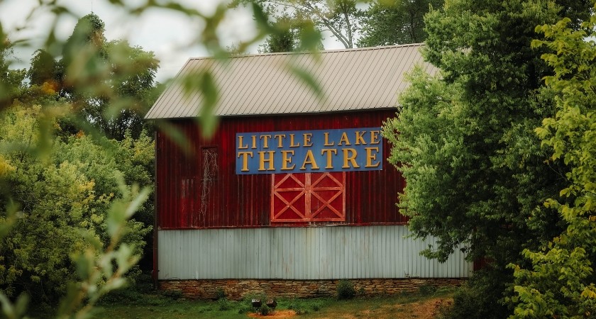 A photo of a barn building through the trees with a sign that says Little Lake Theatre 