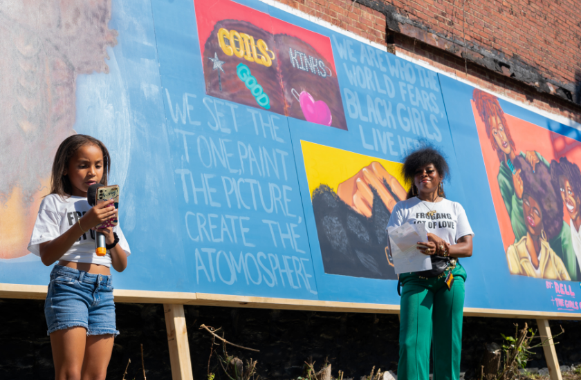 Two people stand in front of a mural featuring artwork and empowering text