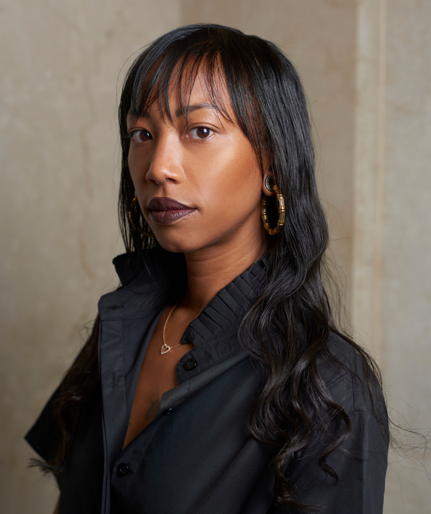 Woman with long dark hair and black shirt standing against a beige background.