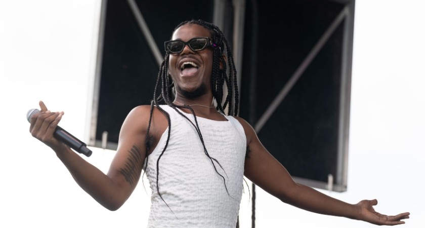 A black man with long black braids and a white tank top smiling and holding a microphone