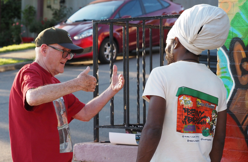 An older man gestures while talking to a man with a turban near a mural.