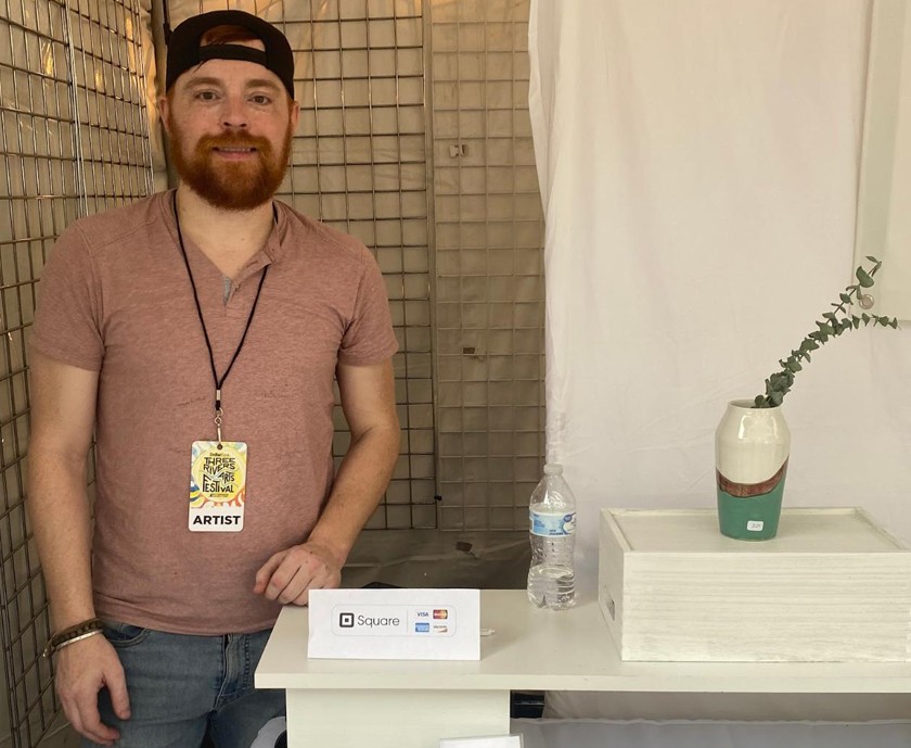White guy with red beard and baseball cap stands in a booth next to a ceramic vase