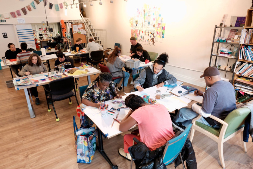 People sitting at tables in a bright art studio, drawing and painting with art supplies.