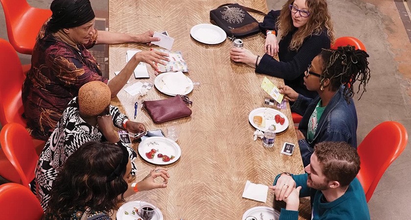 Six racially diverse people sit together in red chairs around a table with plates of food and drink in front of them