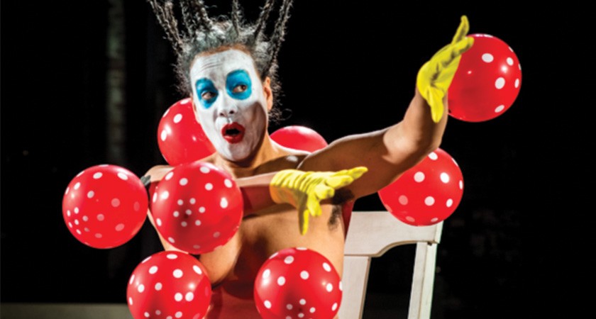 A topless person wearing bright yellow gloves; white, blue, and red face makeup; and whose dark hair is in a bunch of small braids standing up in the air, sits on a white chair while playing with red-and-white balloons