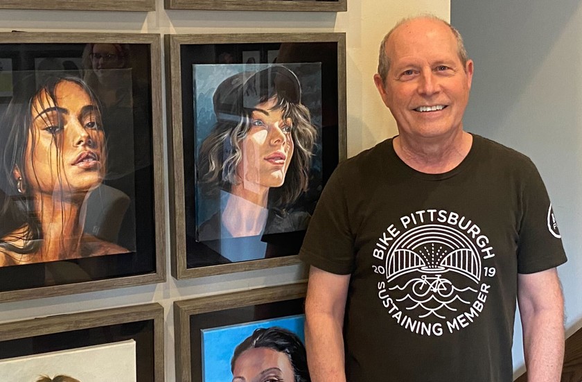 A white man wearing a black Bike Pittsburgh t-shirt stands smiling next to framed acrylic portrait paintings