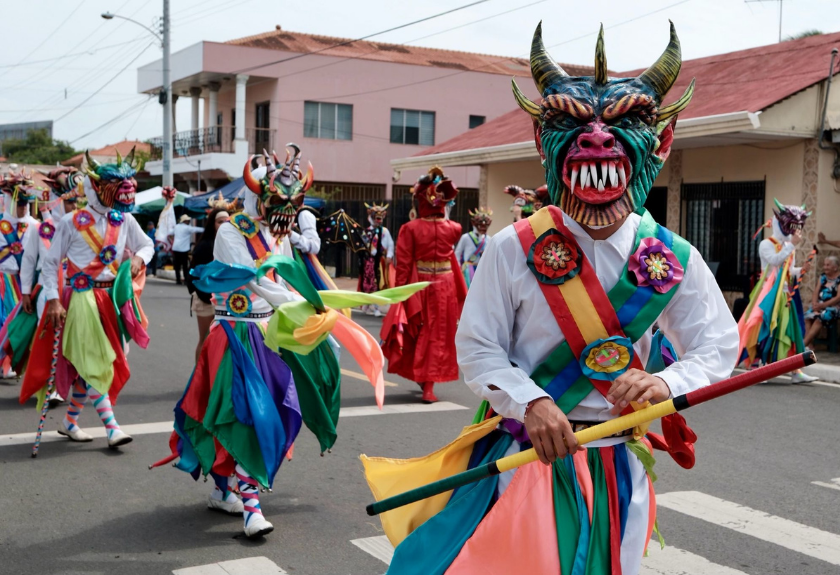 People in colorful demon masks and costumes parade down a street during a festival.