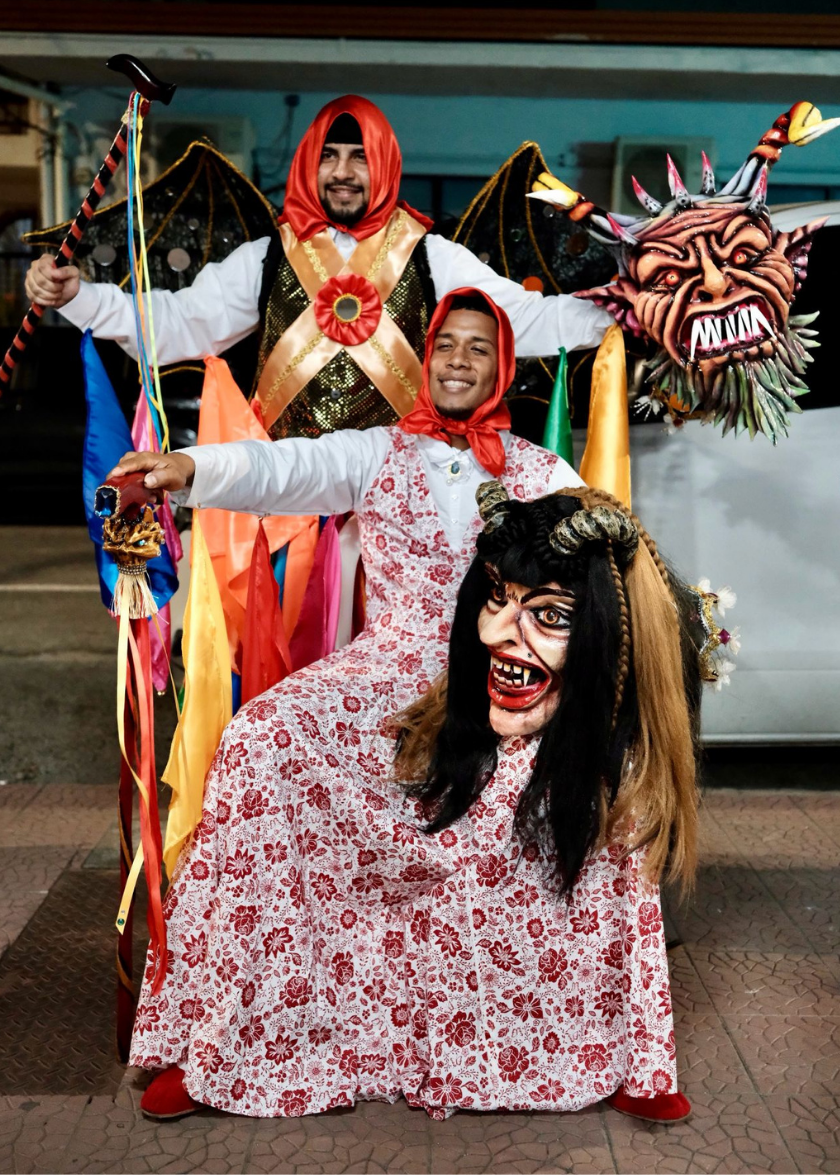 Two people in colorful costumes and masks pose, holding vibrant ribbons and a horned demon mask.
