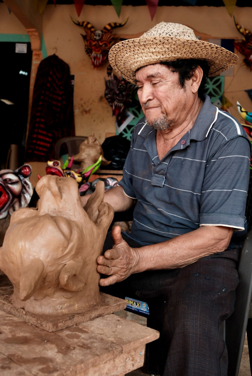 An elderly man shapes a clay sculpture while wearing a straw hat and striped shirt.
