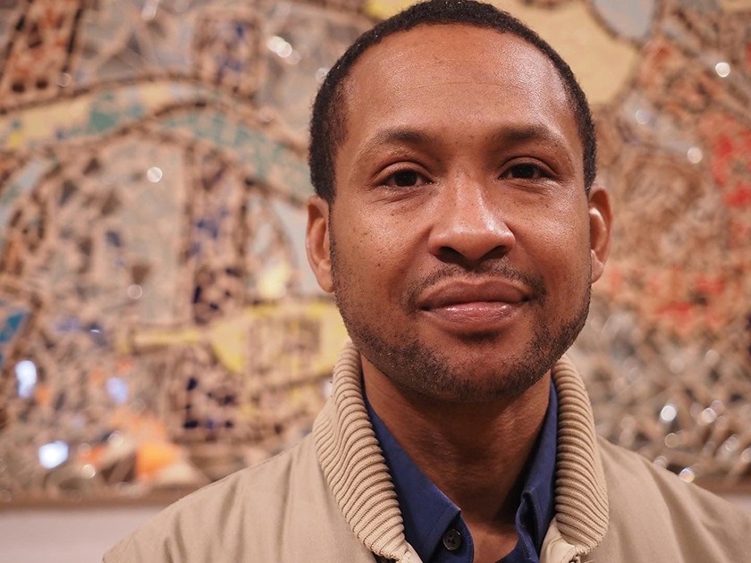 A smiling Black man with short dark hair, a subtle closely-shaved beard, wearing a blue button-down shirt and a tan jacket. Behind him in a colorful tiled artwork