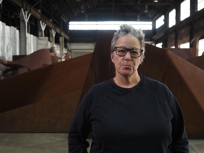 A white woman with short gray hair wears a black sweatshirt and dark glasses. She's slightly smiling and standing in front of a large brown sculpture inside a studio