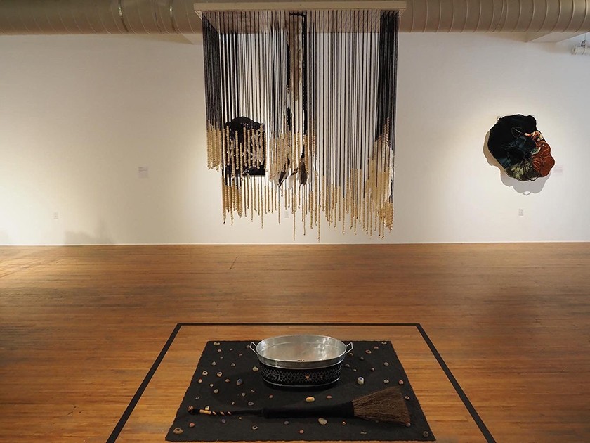 Multiple mixed-media pieces are displayed inside a gallery. On the wooden floor sits a black rug, a broom, and a silver bucket. Above the bucket are beaded strings hanging from the ceiling.