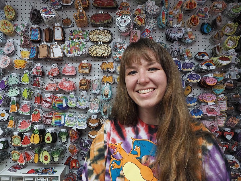 Smiling woman with long dark blonde hair and bangs wearing a tie-dyed shirt with a cartoon flying dragon stands smiling in front of a display full of colorfully illustrated cartoon stickers