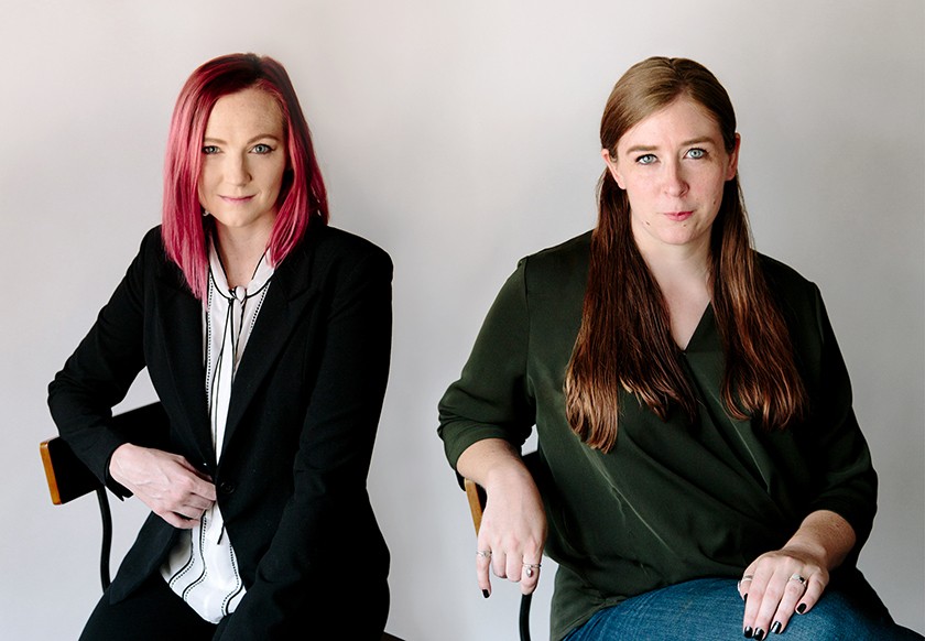 A white woman with shoulder-length straight pink hair, wearing a white button-down shirt and a black blazer, sits in a chair beside a white woman with long straight brown hair, jeans, and a dark green long-sleeved shirt