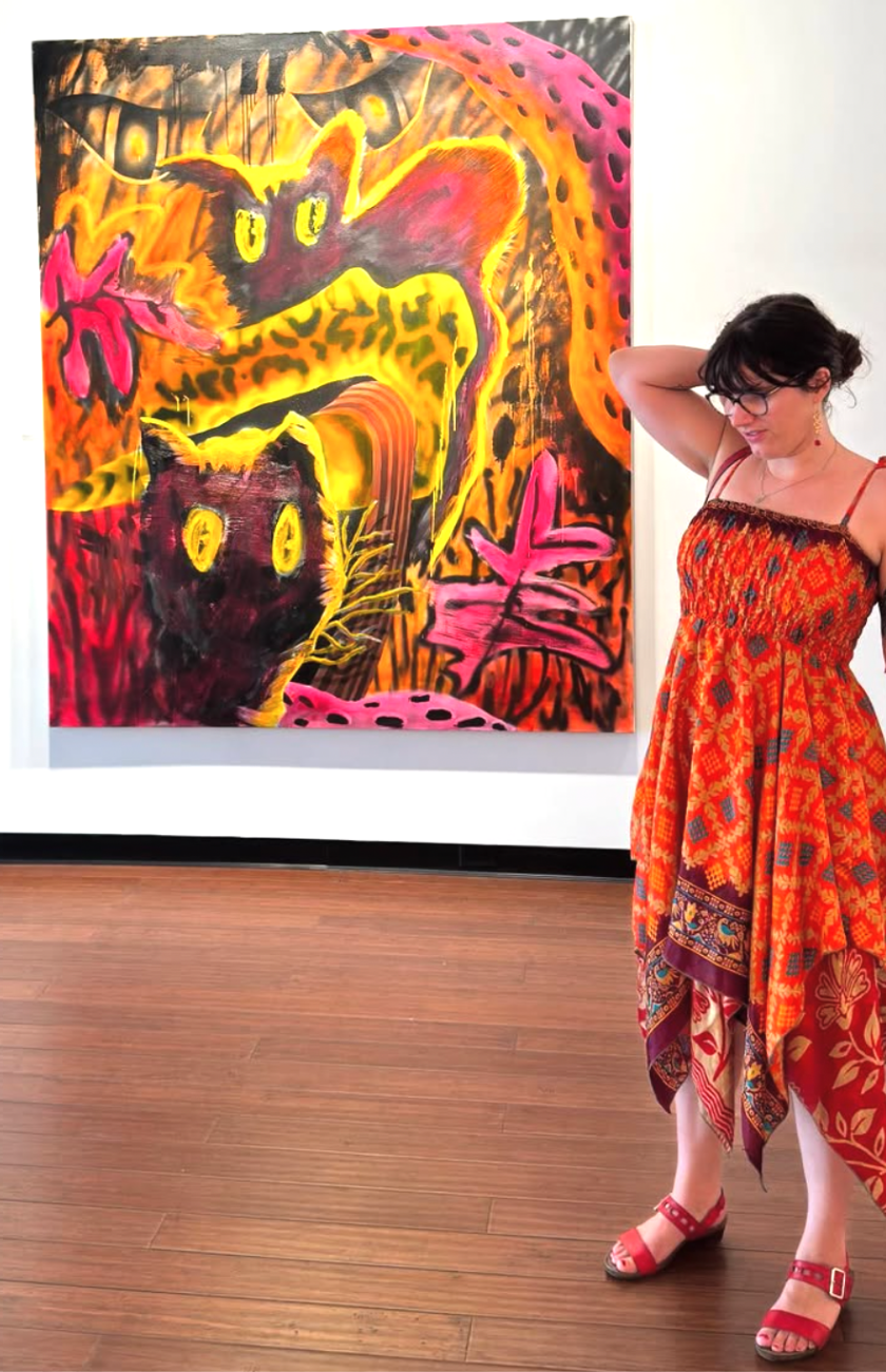 Woman in a red dress stands by a colorful painting of abstract cats in a gallery.