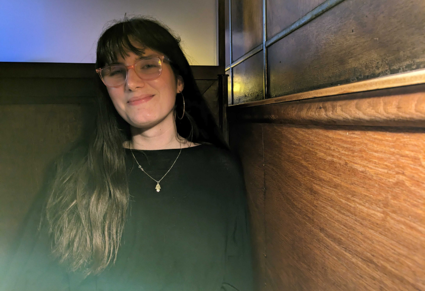 Woman with long dark hair and glasses smiling, sitting by a wooden wall in soft lighting.