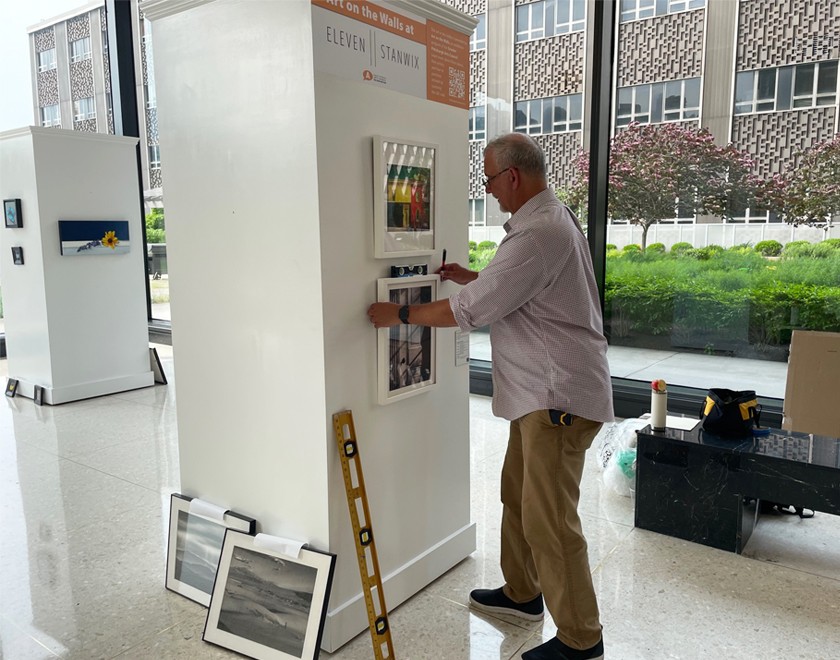 A white man with gray hair and glasses, wearing a button-down dress shirt and a black watch, hangs artwork on a white wall next to a window with green grass and a purple tree