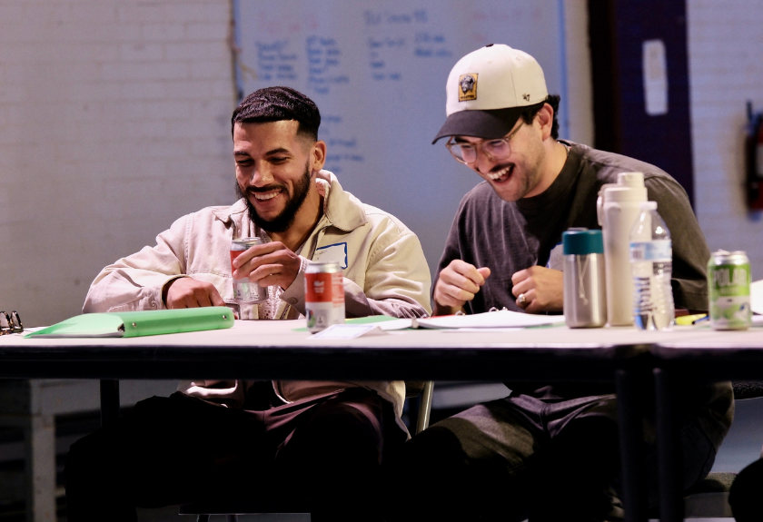 Two men laughing while sitting at a table with beverages and papers.