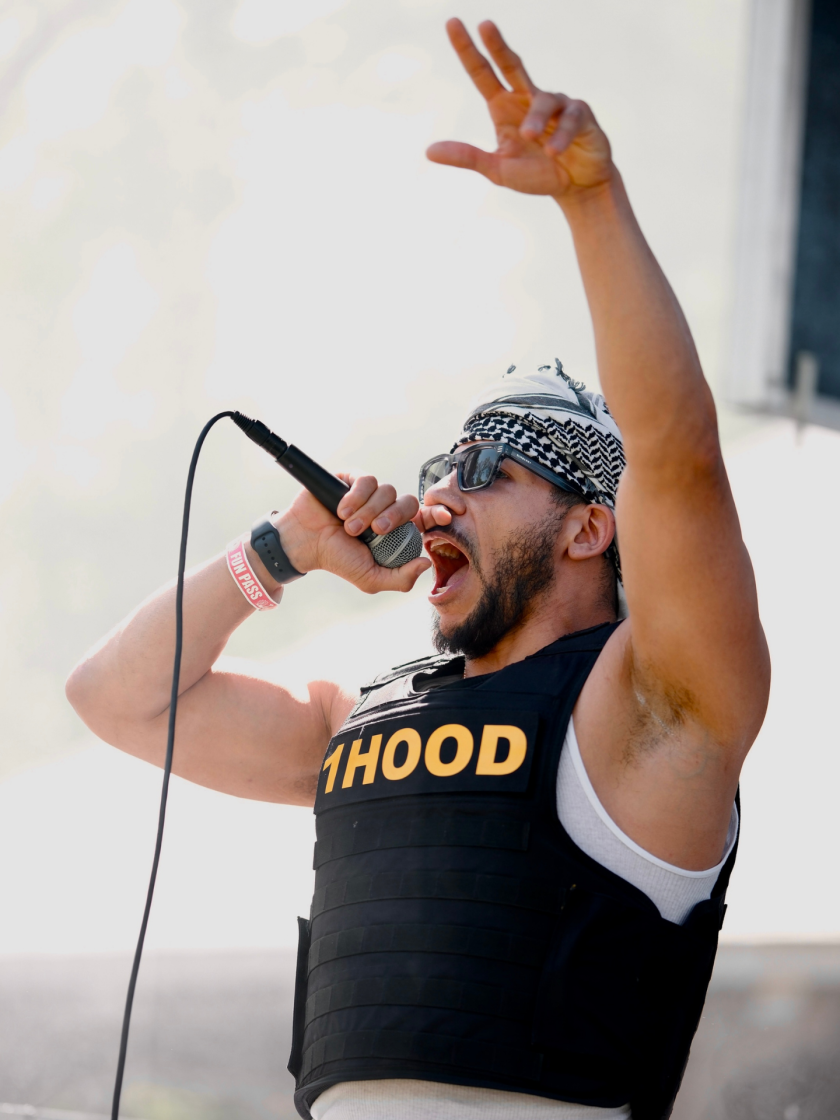 A performer wearing a tactical vest labeled 1HOOD sings into a microphone with one arm raised.