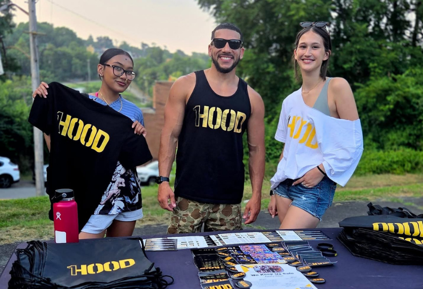 Three people stand behind a table of HOOD branded apparel and materials.
