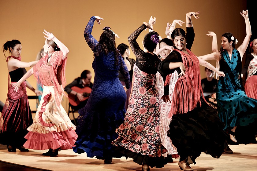 A group of dancers in colorful, traditional dresses and shawls perform a lively flamenco dance on stage, with musicians playing guitar in the background.