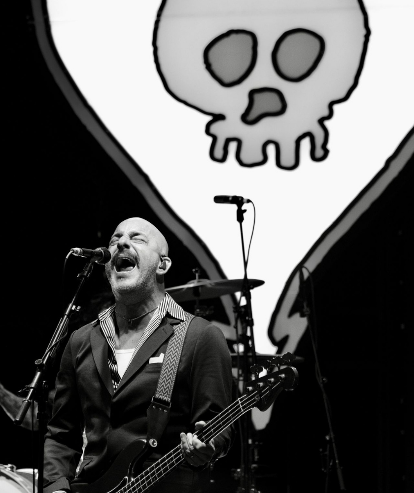 A bald musician passionately sings and plays bass guitar on stage, with a large white cartoon skull and crossbones backdrop behind him. The image is in black and white.
