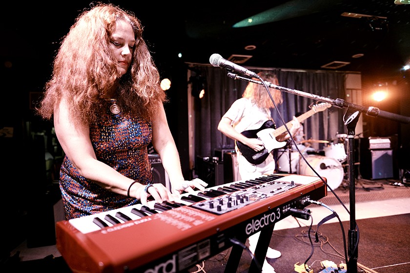 A woman with curly hair plays a red Nord Electro 3 keyboard on stage, while another musician in the background plays an electric guitar. The stage is dimly lit with amplifiers and a drum set visible.