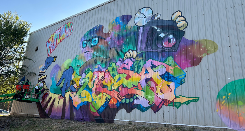 Colorful graffiti mural on a corrugated wall with artists working on a lift