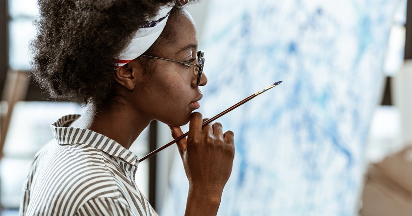 Black woman with short hair pulled back in a headband holds a paintbrush while looking off camera