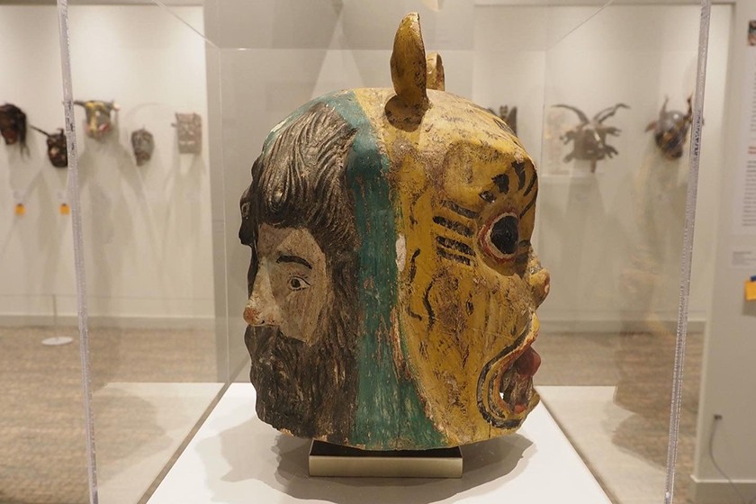 A Mexican mask is displayed inside glass in an exhibition space. There is a face on each side of the mask. On the left side of the mask is a carving of a bearded man. On the right, is an animal with ears and a yellow-and-black painted face
