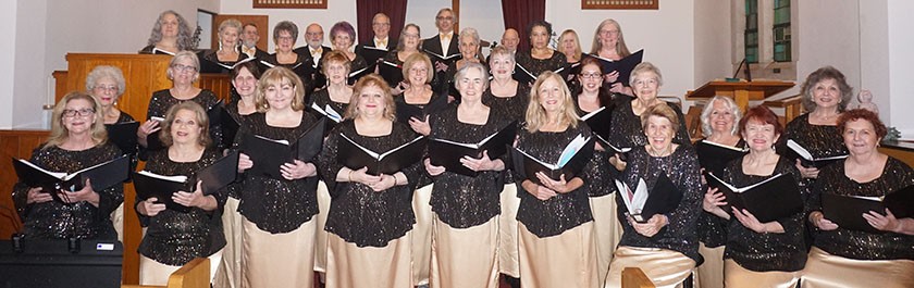 A group of mostly older white women dressed in sparkly black shirts and tan skirts pose smiling while holding choir books