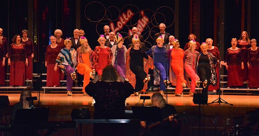 A group of older white singers perform on a stage in front of a conductor and a person playing the guitar