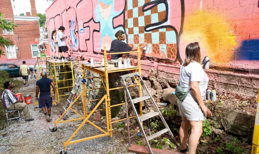 A group of people work together to paint a colorful mural on a large outdoor wall, using ladders and scaffolding. Paint cans and supplies are scattered around as they add new designs to the wall.