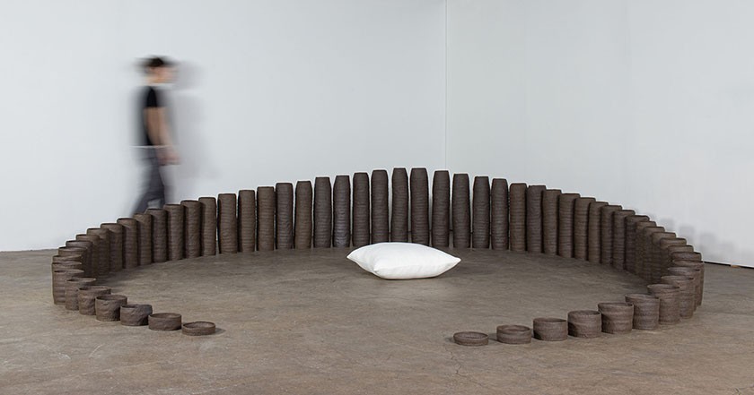 A moving person is photographed in a blur walking behind a row of dark circular bowls in ascending sizes displayed in an arc shape on a museum floor