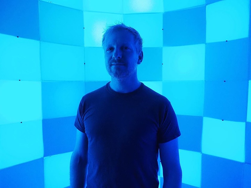 A white man with short light hair and a goatee wearing a dark T-shirt stands in front of a light installation, made up of different colored blue squares. The entire photograph is blue-tinted