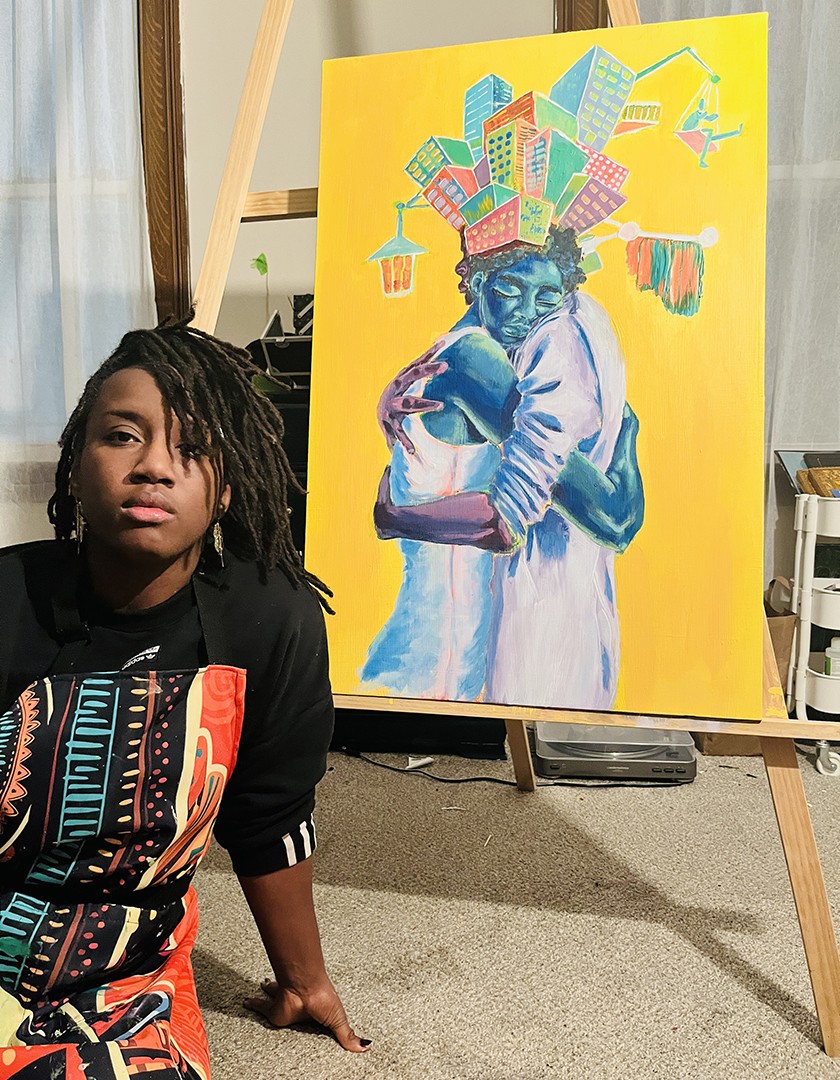 Ifeoma, a Black woman with shoulder-length dark hair wearing a black T-shirt with a colorful Africana-inspired pattern, sits in front of a colorful piece of artwork on display depicting two people hugging on a yellow background