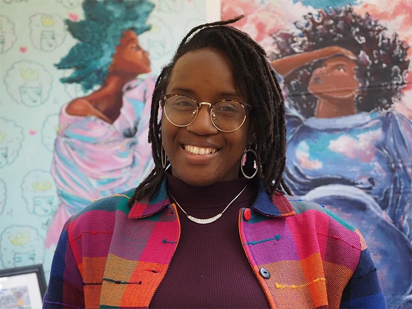 Smiling Black woman with shoulder-length dark hair, gold glasses, hoop earrings, and a colorful plaid shirt. Behind her is colorful artwork of two Black women