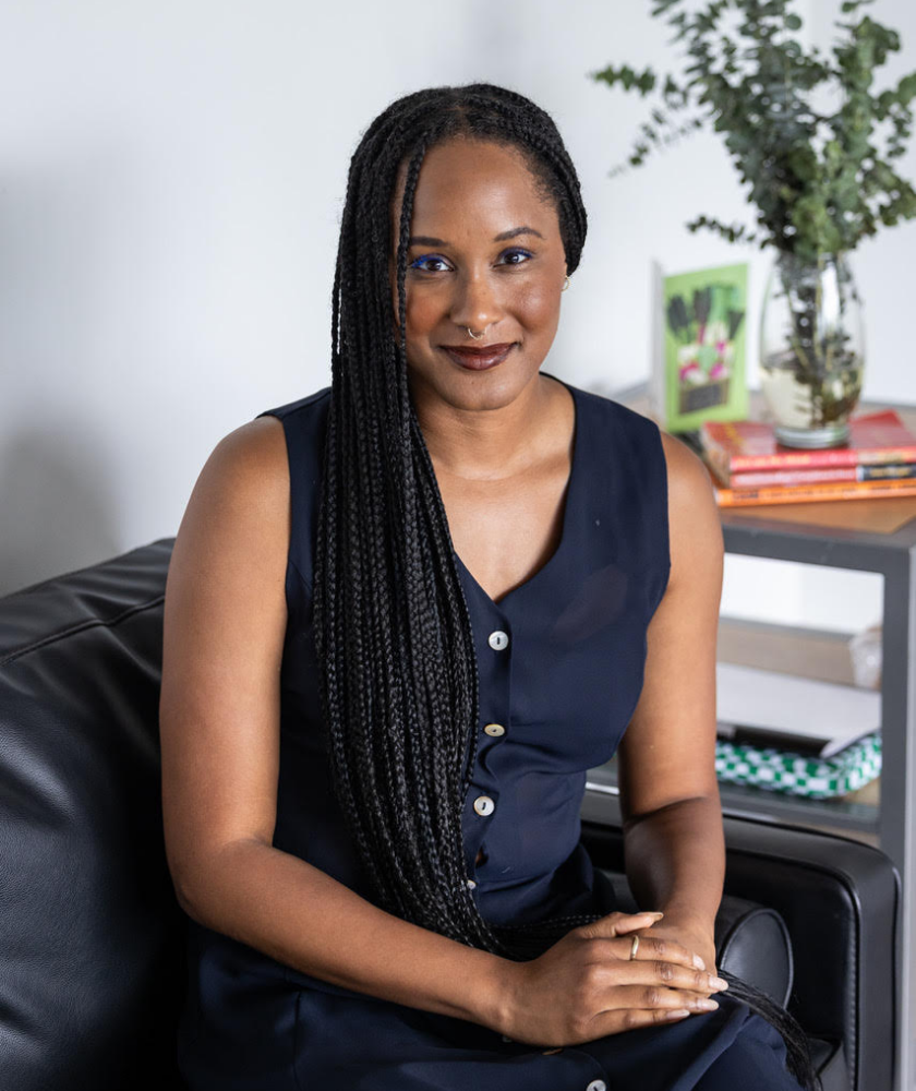 A person with long braided hair, wearing a sleeveless navy dress, sits on a black leather couch. Behind her is a small table with books, a vase of greenery, and a framed photo. She smiles slightly at the camera.