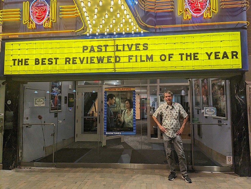 A white man with gray hair, a patterned short-sleeved collared shirt, gray slacks, and sneakers stands outside on the sidewalk in front of a movie theater. The marquee on the theater says, "Past Lives, the best reviewed film of the year"
