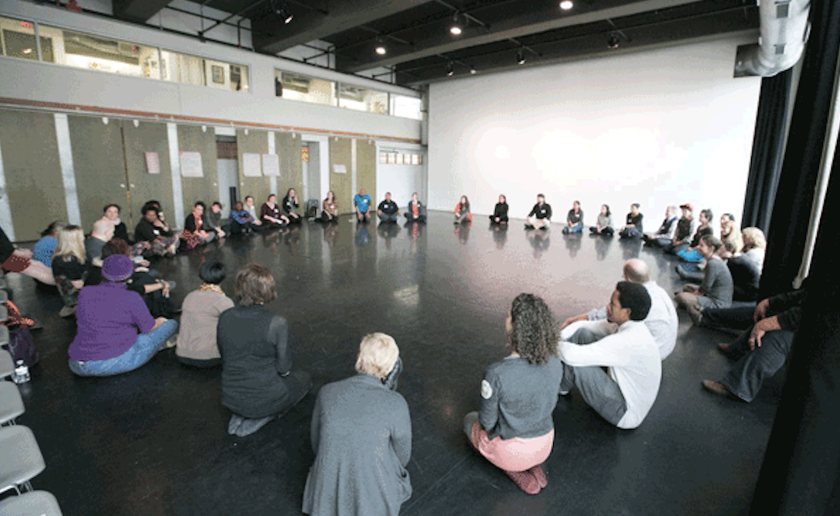 A large group of people sit in a circle on a shiny black floor