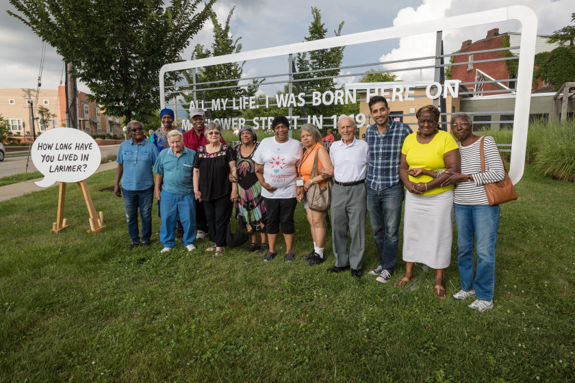 A group of people pose together outside near a small sign of a voice bubble that asks, How long have you lived in Larimer?