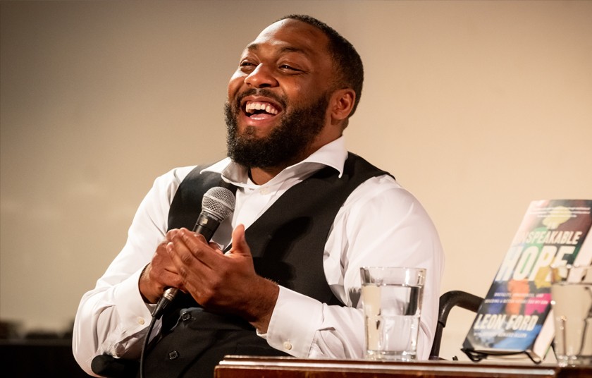 Leon Ford, a Black man with short dark hair and a beard, sits at a table beside his book, holding a microphone and smiling