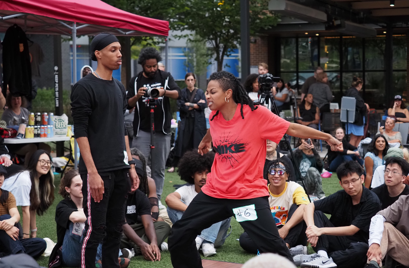 Two dancers face off in an outdoor battle, watched by a crowd seated on the grass.