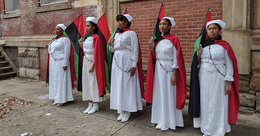 Five people stand in a row waring white gowns, white boots, beaded sashes, white caps, and red capes. All are holding red, black, and green striped flags