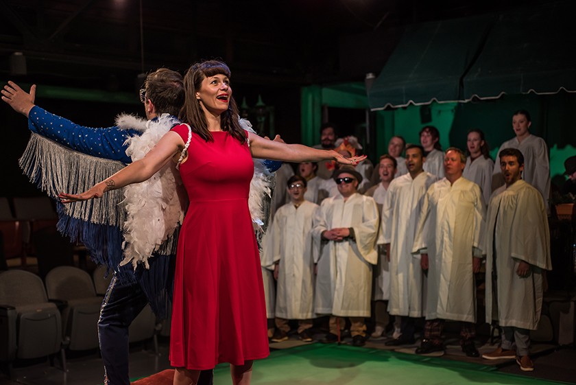 Two actors stand back-to-back on stage. One is a white woman with long brown hair and brown bangs, wearing a red short-sleeved dress, and white angel wings strapped to her arms. Behind her stands what looks like a white man with short brown hair, wearing a blue jacket